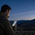 Pria di puncak gunung menggunakan ponsel pintar yang terhubung ke jaringan internet Starlink operator seluler di bawah langit malam.