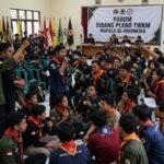 Foto ilustrasi situasi ramai dan penuh interaksi di dalam ruang sidang "Forum Sidang Pleno TWKM Mapala se-Indonesia". Ratusan peserta mahasiswa pecinta alam (Mapala) mengenakan jaket lapangan (PDL) dan slayer merah-oranye duduk berkerumun di lantai serta kursi plastik sambil berdiskusi dengan intens. Tampak spanduk di dinding bertuliskan "FORUM SIDANG PLENO TWKM MAPALA SE-INDONESIA" dengan jajaran bendera organisasi di bagian belakang. Beberapa peserta berdiri sambil mengacungkan tangan dan berbicara menggunakan mikrofon di tengah kerumunan yang padat.